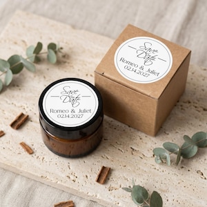 May include: A small, round jar with a black lid and a white label that reads "Save the Date" above "Romeo & Juliet 02.14.2027." A small, square, brown box with a matching label is next to the jar. Cinnamon sticks and greenery are scattered around.