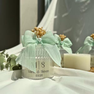 Puede incluir: Un tarro de vela de cristal transparente y estriado con un lazo de tela verde claro y una pequeña cuenta blanca en el centro. La etiqueta muestra las iniciales M | S, los nombres Marie & Steven y la fecha 12.10.26. Flores secas tras el lazo.