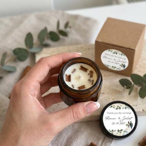 Op de afbeelding: Een kleine, ronde, amberkleurige glazen kaarsenpot gevuld met witte was en kaneelstokjes. De kaars wordt in een hand gehouden. Een kleine bruine doos en een kaarsdeksel met tekst en bloemmotief zijn ook zichtbaar.