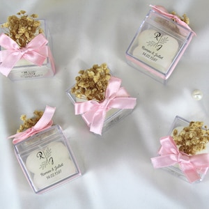 May include: Four clear square boxes with pink bows and dried flowers. Each box contains a white round favour with the text "R & J Romeo & Juliet 14.02.1597".