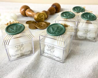 Puede incluir: Cajas cuadradas transparentes atadas con cinta dorada y blanca, cada una coronada con un sello de cera verde. Las cajas contienen objetos blancos y texto impreso. Un sello de madera y sellos de cera dorada están en el fondo. Recuerdos de boda.