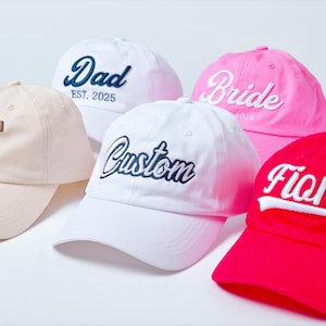 Könnte beinhalten: Eine Auswahl an Baseballkappen in verschiedenen Farben, darunter Weiß, Rosa und Rot. Jede Kappe hat einen individuellen gestickten Text, wie "Dad", "Bride", "Custom", "Fiona" und Initialen. Die Kappen sind auf einer weißen Oberfläche ausgestellt.