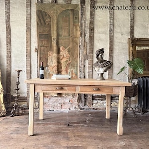 May include: A light-coloured wooden table with two drawers, a wine bottle, and a bust of a person. The table is in a room with antique decor, including a large painting and ornate mirrors. The room has a rustic, vintage feel.