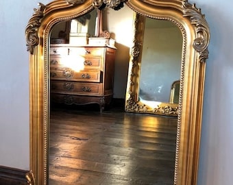 May include: A large, ornate gold-framed mirror with a beveled edge and intricate carvings. The mirror is arched at the top and has a decorative design around the perimeter.