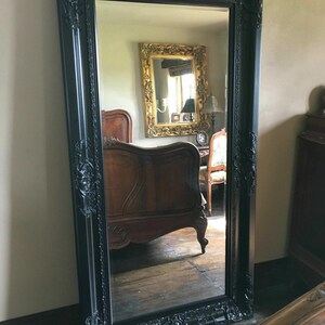 May include: A large rectangular floor mirror featuring a thick black frame with ornate, raised scrollwork and floral carvings along the edges. The mirror surface reflects a room containing a wooden bed frame and a smaller gold-framed mirror.