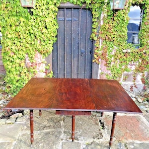 May include: A mahogany drop leaf table with four turned legs. The table is set up outdoors on a stone patio with a brick wall and ivy covered wall in the background. The table is extended with two drop leaves.