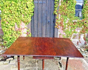 May include: A mahogany drop leaf table with four turned legs. The table is set up outdoors on a stone patio with a brick wall and ivy covered wall in the background. The table is extended with two drop leaves.