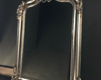 May include: A silver ornate mirror with a decorative, ornate frame. The frame has a raised, detailed design with a floral motif. The mirror is rectangular with an arched top.