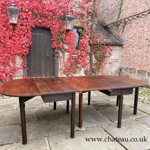 May include: A large, oval wooden table with a dark finish and two drawers. The table is supported by dark, slender legs. The background features a brick building and a door covered in red foliage. The website address www.chateau.co.uk is visible.