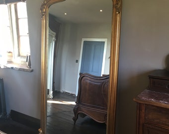 May include: A tall, ornate gold-framed mirror with a decorative top. The mirror is reflecting a room with a wooden floor and a dark wooden chair.
