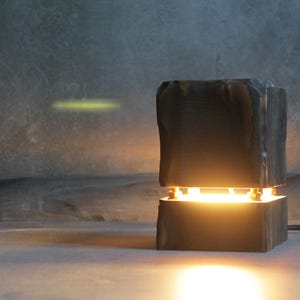 May include: A contemporary, dark grey wooden table lamp emitting a warm, yellow light. The lamp features a textured, blocky design and is placed on a grey surface. A black power cord extends from the base.