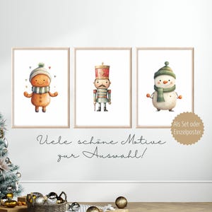 May include: Three framed wall art prints featuring watercolor illustrations of a gingerbread person, a nutcracker, and a snowman, each wearing a green and white striped scarf and hat, displayed on a white wall with text in German.