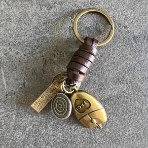 Può includere: Un portachiavi in ​​cuoio marrone con un ciondolo in metallo dorato che dice "LOVE" e un piccolo ciondolo rotondo in metallo dorato con un design circolare. Il portachiavi ha anche un piccolo ciondolo rettangolare in metallo dorato con il testo "It is about you, makes my heart long to be free".