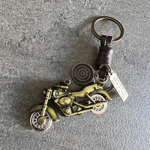 Op de afbeelding: Een gouden motorfiets sleutelhanger met een bruin leren bandje en een metalen ring. De motorfiets is gedetailleerd ontworpen en de sleutelhanger bevat een bedel met de tekst "I feel about you makes my heart long to be free".