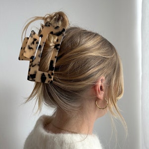 Op de afbeelding: Een grote schildpadkleurige haarspeld met een rechthoekige vorm. De speld houdt het blonde haar van een vrouw in een knot.
