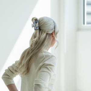 Può includere: Una persona con i capelli chiari indossa un maglione color crema con una clip per capelli a fantasia blu e bianca. La clip per capelli tiene i capelli in uno chignon.