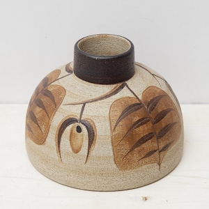 May include: A ceramic vase with a wide, rounded base and a dark brown rim. The beige surface is decorated with brown leaf and stem designs. The vase has a rustic, handcrafted appearance, suitable for home decor.