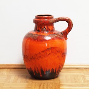 Op de afbeelding: Een grote, oranje keramische vaas met een handvat. De vaas heeft een glanzende afwerking met donkerbruine en zwarte accenten. Het handvat is gebogen en bevestigd aan de bovenkant van de vaas. De vaas is ongeveer 30 cm hoog.