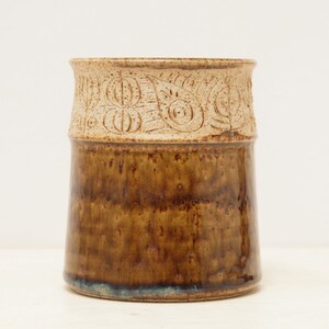 Puede incluir: Un jarrón de cerámica marrón con una banda blanca alrededor de la parte superior. La banda tiene un diseño tallado de hojas y círculos.