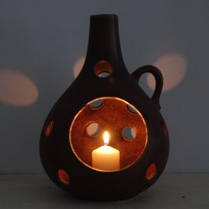 May include: A dark brown ceramic candle holder with a handle and multiple circular cutouts. A lit white candle is inside, casting a warm glow through the openings. The candle holder has a teardrop shape.