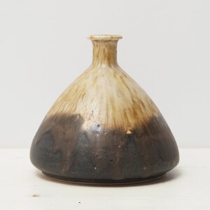 Könnte beinhalten: Eine handgefertigte Keramikvase mit einer einzigartigen, organischen Form. Die Vase weist einen Farbverlauf auf, der von hellem Beige oben nach tiefem Braun unten übergeht. Die Vase ist etwa 15 cm hoch.