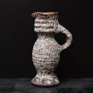 Può includere: Brocca in ceramica con manico, caratterizzata da un design bianco e marrone maculato. La brocca ha una superficie strutturata e un'estetica rustica unica. Il bordo e la base sono di un marrone più scuro.