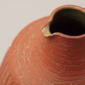 Puede incluir: Primer plano de la parte superior de una jarra de terracota con una abertura ancha y redondeada. La jarra tiene una superficie rugosa y texturizada con l&iacute;neas visibles y un color marr&oacute;n rojizo.