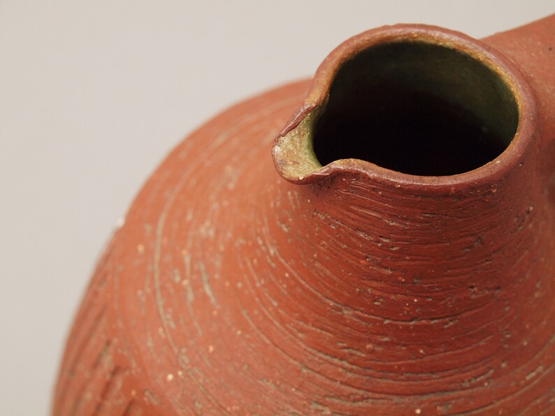 Puede incluir: Primer plano de la parte superior de una jarra de terracota con una abertura ancha y redondeada. La jarra tiene una superficie rugosa y texturizada con l&iacute;neas visibles y un color marr&oacute;n rojizo.
