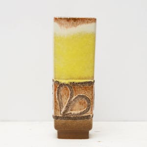 May include: A tall, rectangular ceramic vase with a gradient of colours. The top is brown and white, transitioning to yellow, with a textured brown base featuring a heart-shaped design. The vase is approximately 30 cm tall.