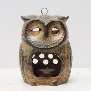 May include: A ceramic owl-shaped candle holder in shades of brown and tan. The owl has large eye openings and a circular opening in the front with several smaller holes. A small candle sits inside.