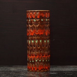 May include: A tall, cylindrical ceramic vase with a textured surface. The vase has a repeating geometric pattern in brown, orange, and green. This decorative vase is suitable for displaying flowers or as a standalone art piece, and is a stylish home decor item.