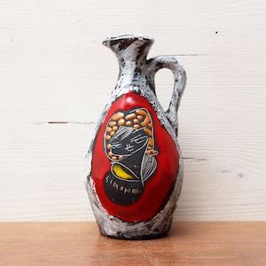 May include: A ceramic pitcher with a white glaze and a red, black, and yellow design. The design features a stylized face with a yellow halo and the word "LIMONIER" written in black.
