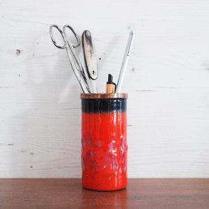 May include: A red and black ceramic desk tidy with a copper rim, containing scissors, a pen, and other stationery. The organiser has a textured surface and is on a wooden table, with a white wooden backdrop.