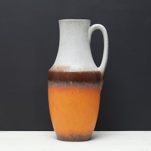 May include: A tall, ceramic pitcher with a handle. The top is white, transitioning to a brown band, and then to a vibrant orange base. The pitcher has a textured surface and a curved handle.