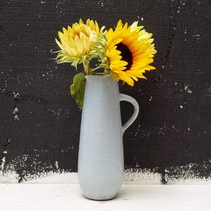 Puede incluir: Una jarra de cerámica azul claro contiene dos girasoles con pétalos de color amarillo brillante y centros de color marrón oscuro. La jarra tiene un asa y se encuentra sobre una superficie blanca contra una pared negra texturizada.