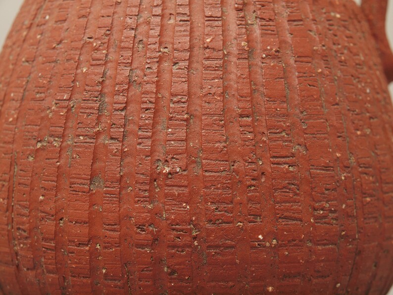 Puede incluir: Primer plano de una maceta de terracota con una superficie texturizada. La maceta es de color marr&oacute;n rojizo y est&aacute; decorada con l&iacute;neas verticales.