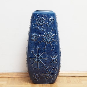 May include: A dark blue ceramic vase with a textured surface. The vase features raised starburst patterns and small, round details. It has a tapered shape and a wide opening. The vase is suitable for floral arrangements or decorative display, and is approximately 30 cm tall.