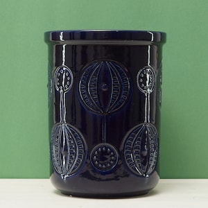 May include: A dark blue ceramic vase with a cylindrical shape and an embossed floral pattern. The vase has a glossy finish and a slightly flared rim. The background is a solid green. The vase is approximately 20 cm tall.