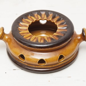 May include: A vintage ceramic fondue pot in shades of brown and tan. The pot features a circular design with a heart-shaped opening in the center and three small holes on the side. It has two handles with a spiral design.