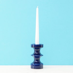 May include: A dark blue ceramic candlestick holder with a white taper candle. The candlestick has a tiered design and a glossy finish. The candle is long and slender. The background is a light blue.