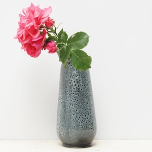 Può includere: Un vaso di ceramica grigio con una superficie strutturata contiene un bouquet di rose rosa e foglie verdi. Il vaso ha un collo stretto e una base più ampia. Le rose sono in piena fioritura e le foglie sono di un verde vibrante.
