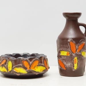 May include: A brown ceramic ashtray and vase set. The ashtray is round with a scalloped edge, and the vase has a handle. Both pieces are decorated with orange and yellow floral designs.