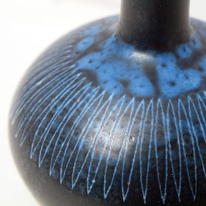 Può includere: Un vaso di ceramica blu scuro con corpo rotondo e collo stretto. Il vaso presenta una superficie strutturata con un motivo di linee bianche curve e accenti blu. È un oggetto decorativo.