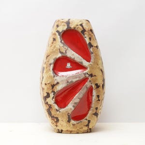 Puede incluir: Un jarrón de cerámica con un exterior texturizado beige y marrón. El jarrón presenta tres grandes aberturas rojas brillantes en forma de lágrima. Un pequeño emblema es visible dentro de una de las formas rojas. Este jarrón decorativo es un artículo de decoración único.