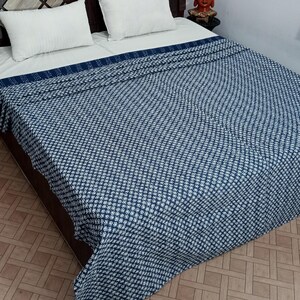 Op de afbeelding: Een bed met een marineblauwe en witte sprei. Twee witte kussens liggen op het bed, dat een donkerbruin hoofdbord heeft. Links staat een rode vaas met gele bloemen.