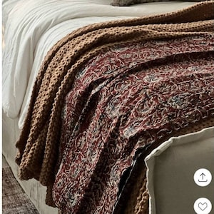 May include: A bed featuring a white duvet, a thick tan knitted throw blanket, and a dark red patterned quilt with intricate floral motifs. The bed is accented with three decorative pillows in plaid, striped, and distressed dark grey patterns.