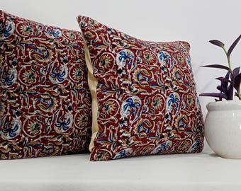 May include: Two square throw pillows featuring a deep red background with a repeating pattern of blue, white, and tan floral and leaf motifs. The pillows have a visible cream-colored side panel and are displayed next to a small white potted plant.