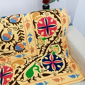 May include: A decorative, embroidered textile with a light yellow background. The textile features a repeating pattern of floral and paisley designs in shades of pink, blue, green, and brown. The textile is draped over a white sofa.