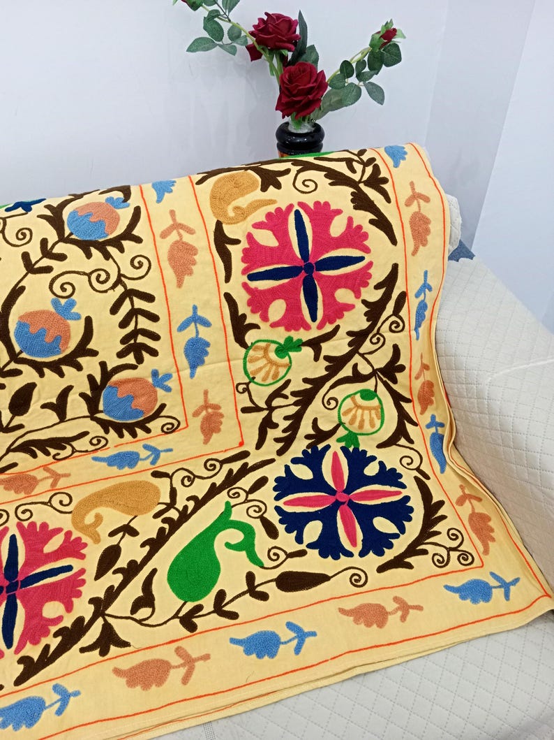May include: A decorative, embroidered textile with a light yellow background. The textile features a repeating pattern of floral and paisley designs in shades of pink, blue, green, and brown. The textile is draped over a white sofa.