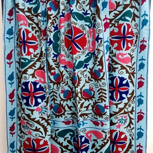 May include: A light blue textile with intricate embroidery. The design features red, blue, pink, and green floral and paisley motifs on a light blue background, with brown vines and a blue border. The textile is likely a decorative throw or blanket.